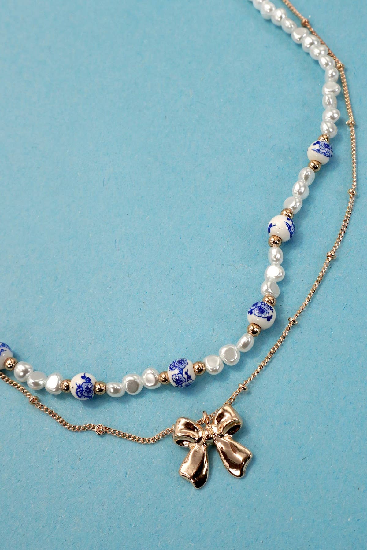 Layered necklace with gold chain with bow charm and white beads with blue toile pattern