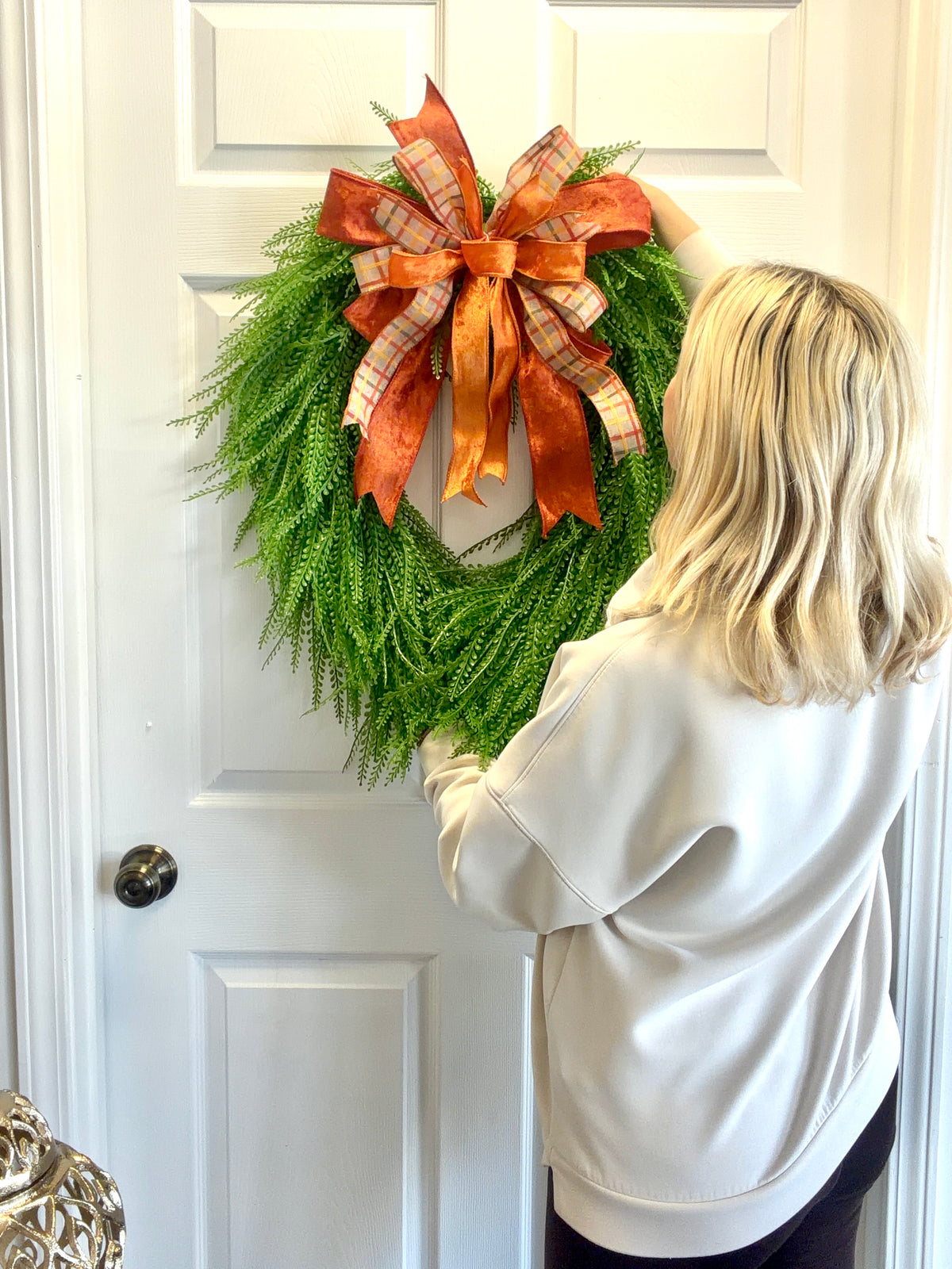 A woman with blonde hair, seen from behind, hanging a green bead grass wreath with a large orange and plaid bow on a white paneled door.