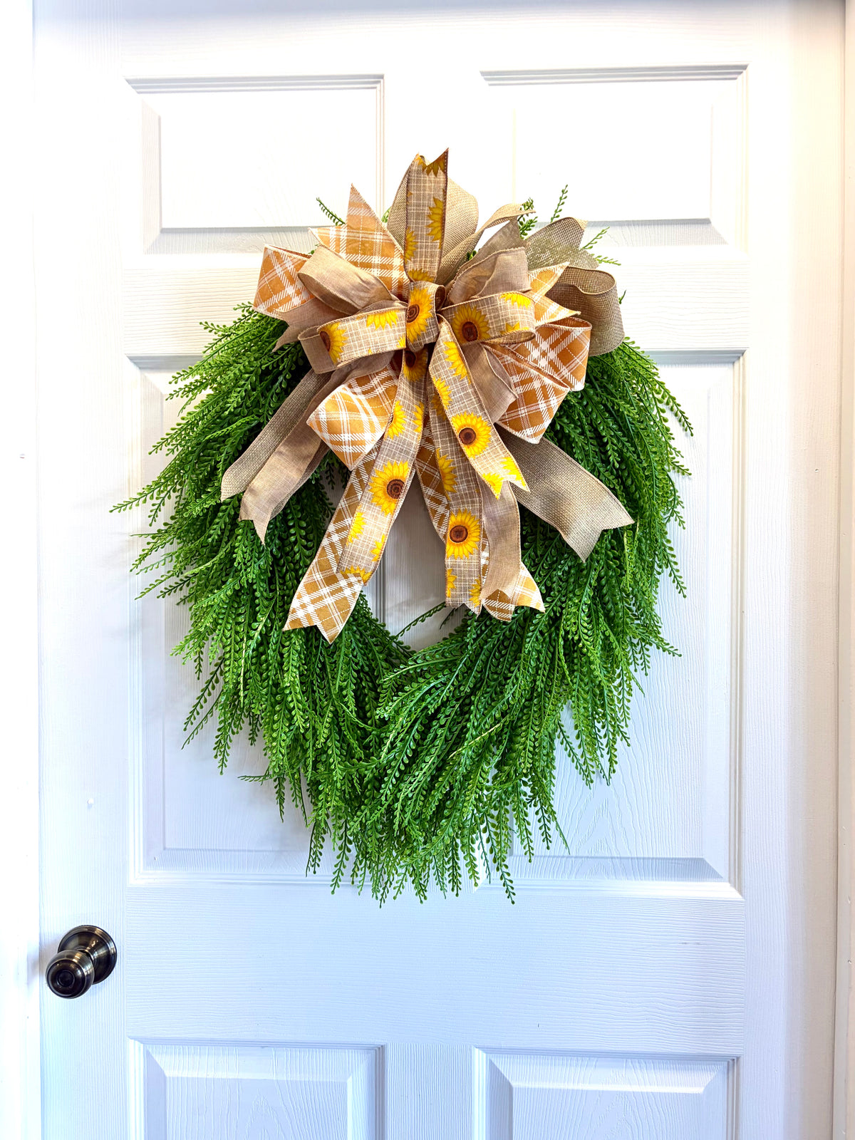 A green beaded fern wreath hanging on a white paneled door, decorated with a large bow made of tan burlap and yellow sunflower-patterned ribbon.