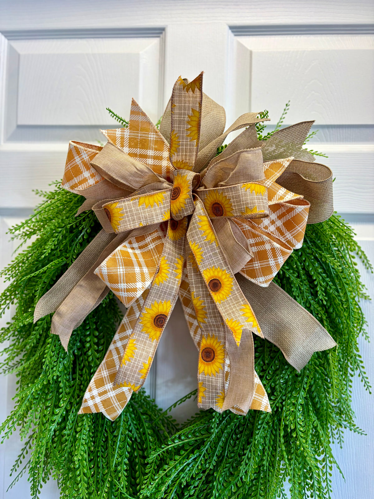 A large autumn-themed bow made from burlap ribbon with yellow sunflower prints and orange plaid patterns, tied around a green artificial wreath in front of a white paneled door.