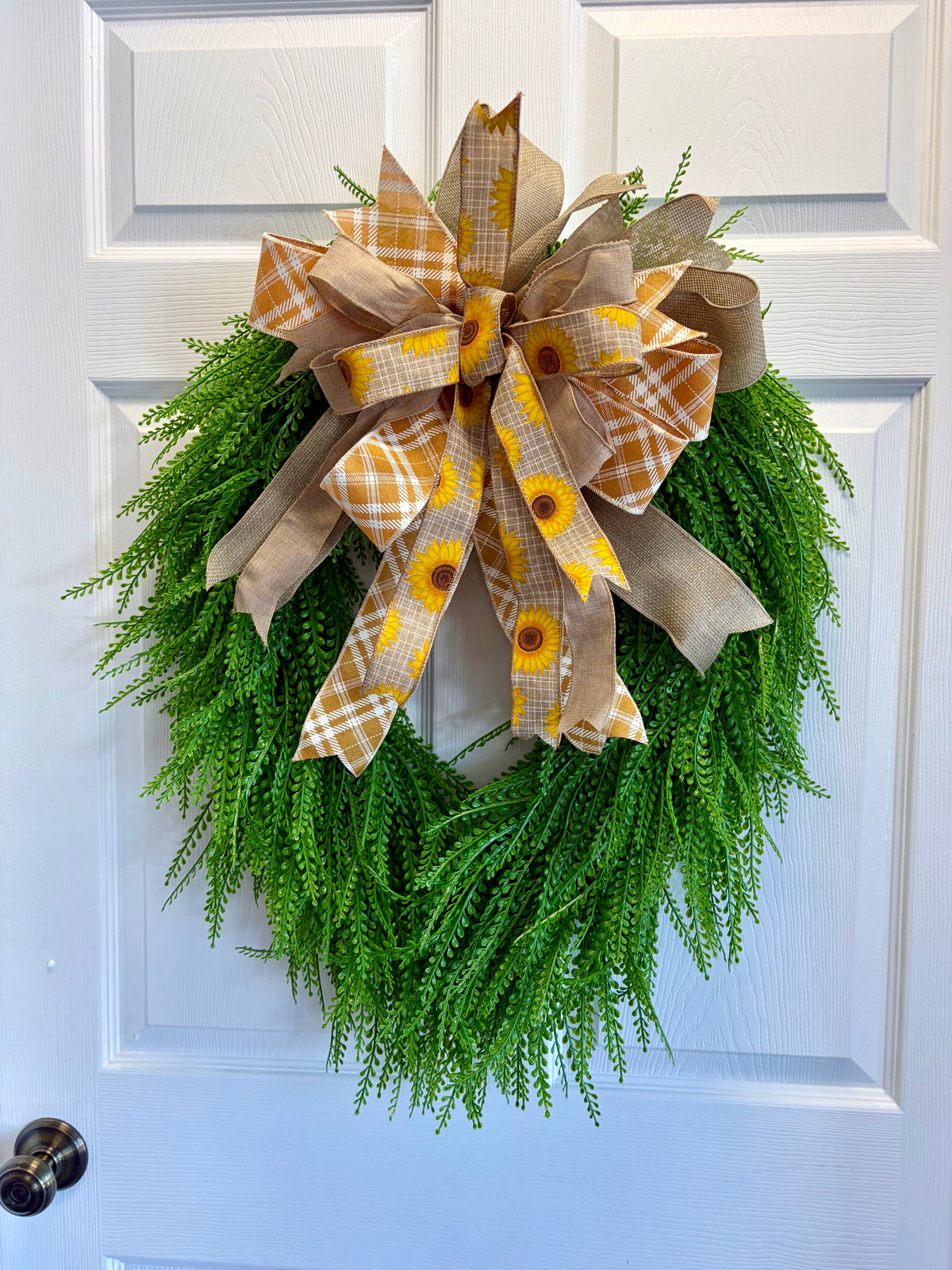 A green artificial fern wreath hanging on a white paneled door, decorated with a large bow made of tan burlap and yellow sunflower-patterned ribbon.