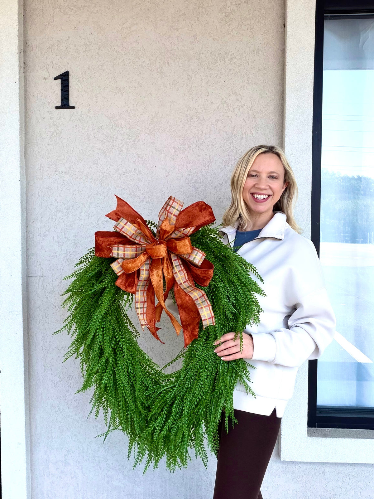 A smiling woman stands outdoors holding a large green wreath decorated with a large orange and brown plaid bow. The background shows a beige stucco wall with a black number '1' and a window.