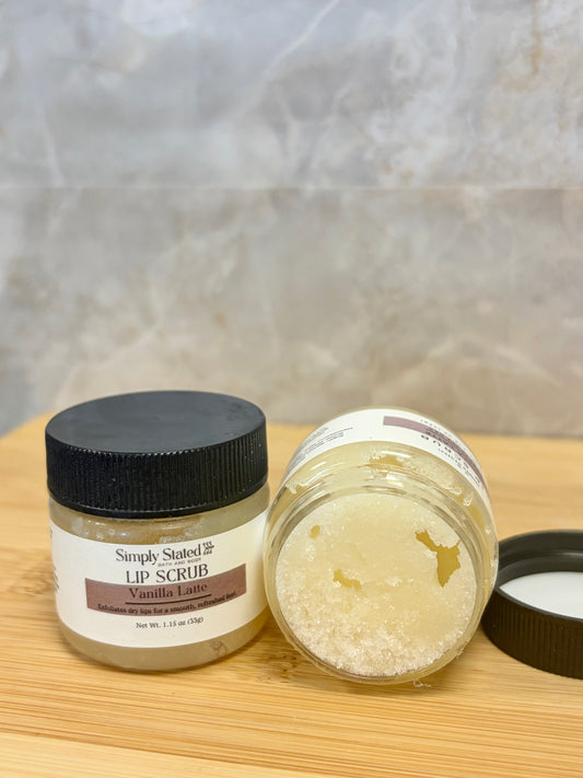 Two small jars of lip scrub on a wooden surface against a gray stone-like background. One jar is closed with a black lid and shows a cream-colored label reading 'Simply Soaked Lip Scrub.