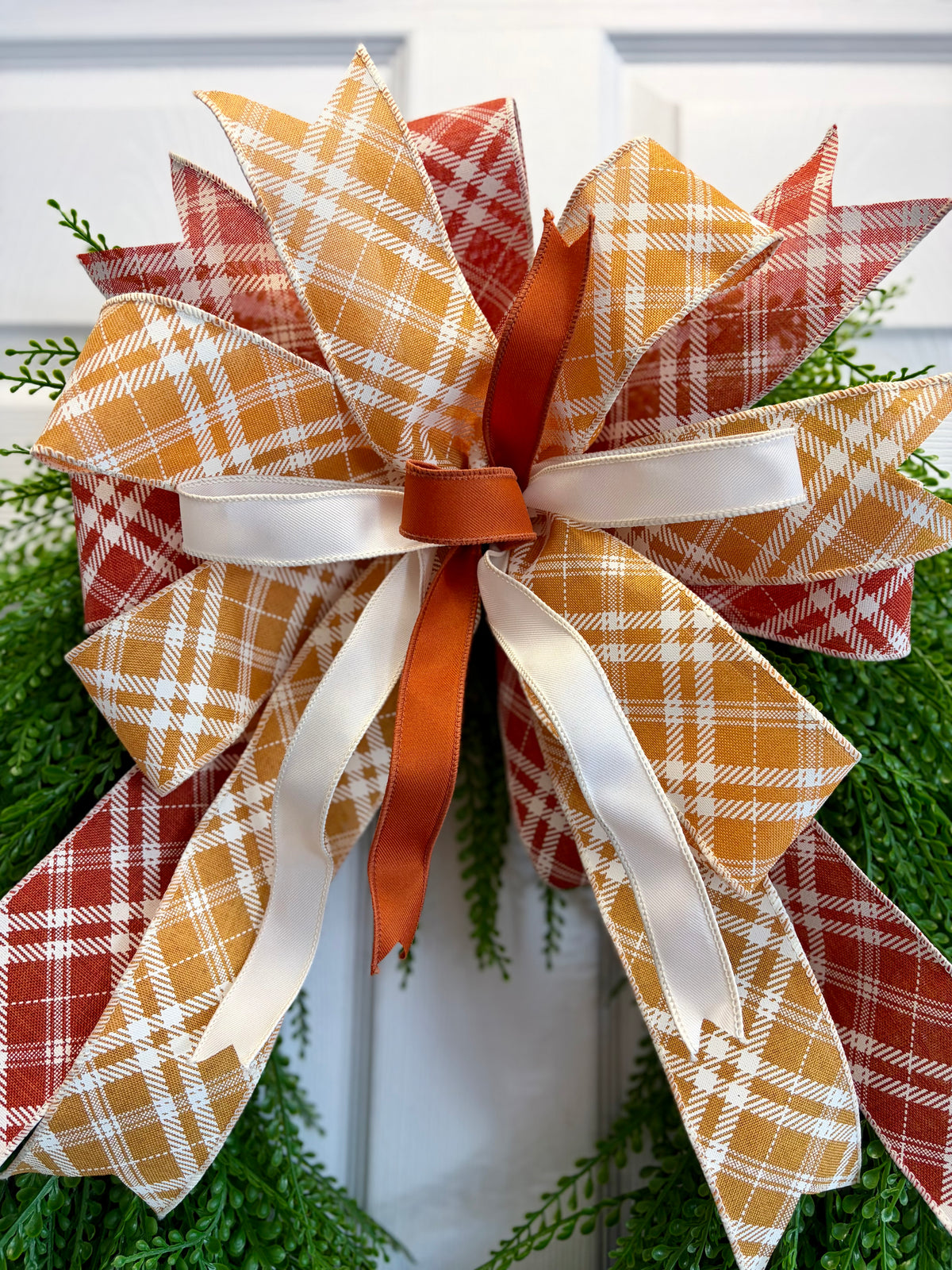 A large autumn-themed decorative bow made from layered plaid and solid ribbons, attached to a green wreath. The bow features orange, rust red, mustard yellow, and white plaid patterns, with a central orange ribbon and white satin ribbon accents.