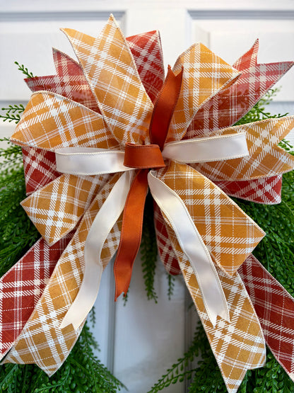 A large autumn-themed decorative bow made from layered plaid and solid ribbons, attached to a green wreath. The bow features orange, rust red, mustard yellow, and white plaid patterns, with a central orange ribbon and white satin ribbon accents.