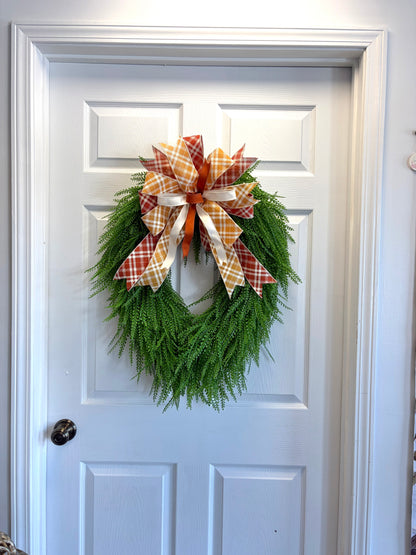 A green bead grass wreath hanging on a white paneled door, decorated with a large orange and white plaid bow.