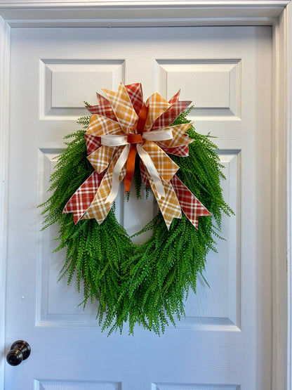 A green artificial fern wreath hanging on a white paneled door, decorated with a large plaid bow in orange, white, and red.
