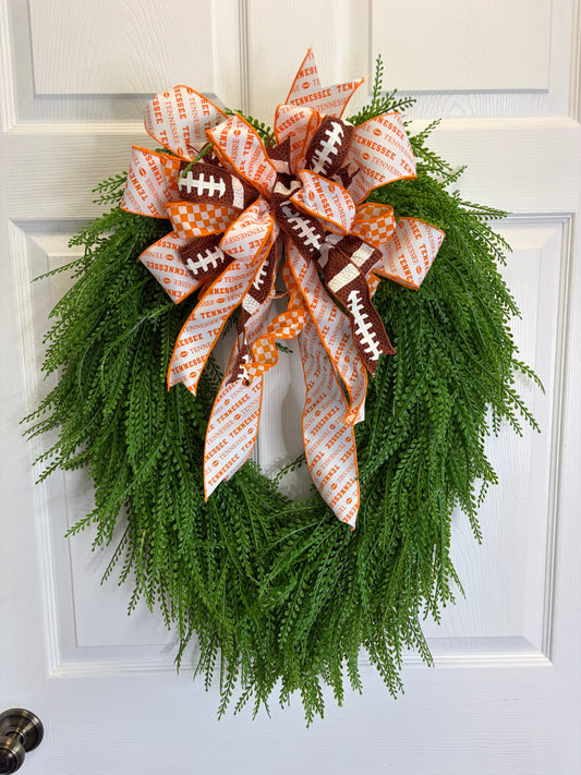 A green artificial fern wreath hanging on a white paneled door, decorated with a large bow made from orange-and-white patterned ribbon, Tennessee ribbon, white-and-orange checkered ribbon, and football-shaped accents.