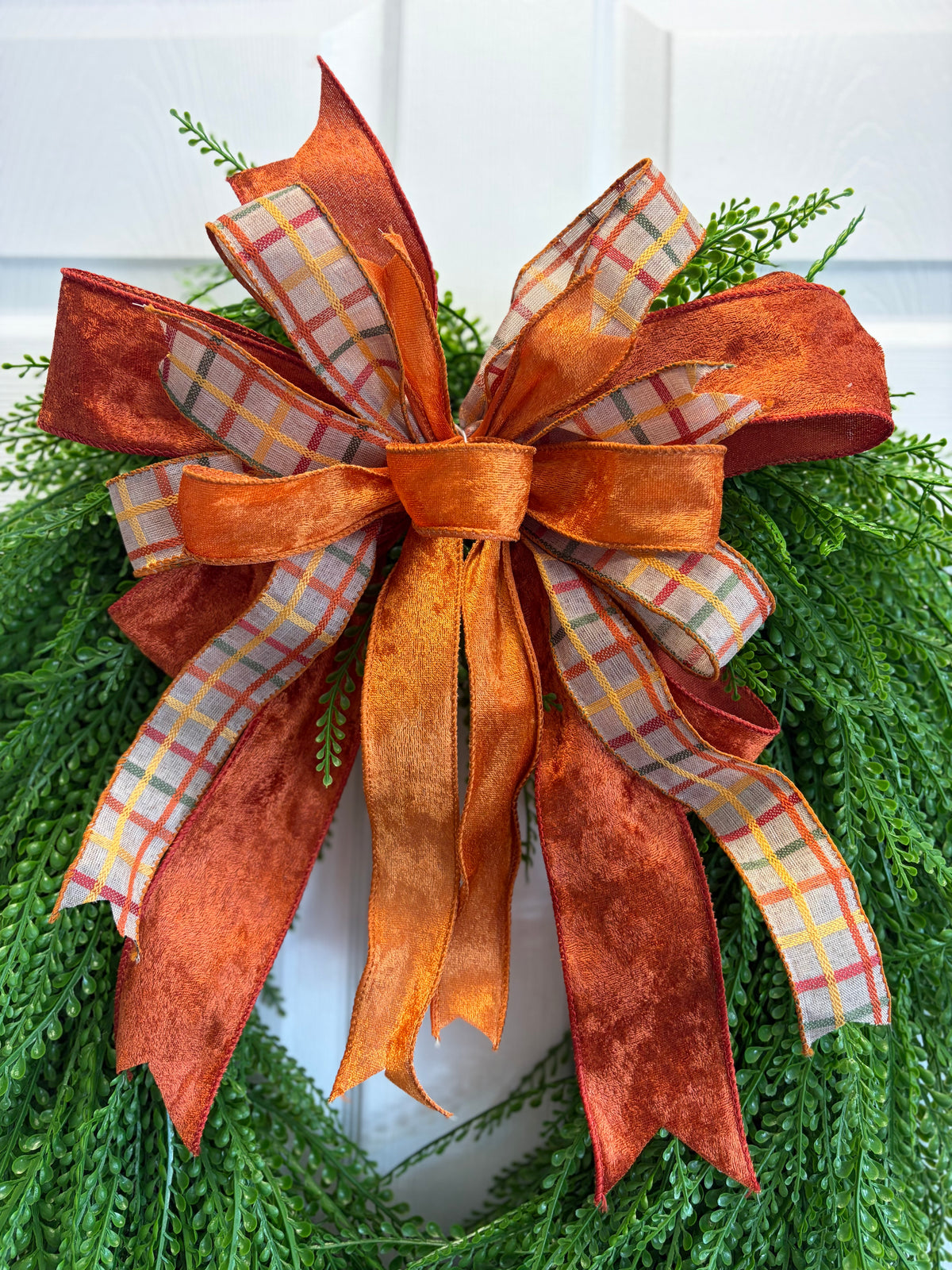 An autumn-themed decorative bow made of layered orange velvety  ribbon, rust-colored fabric, and plaid patterned ribbon, tied on a green pine wreath against a white door background.