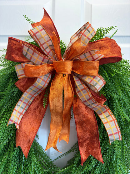 An autumn-themed decorative bow made of layered orange velvety  ribbon, rust-colored fabric, and plaid patterned ribbon, tied on a green pine wreath against a white door background.
