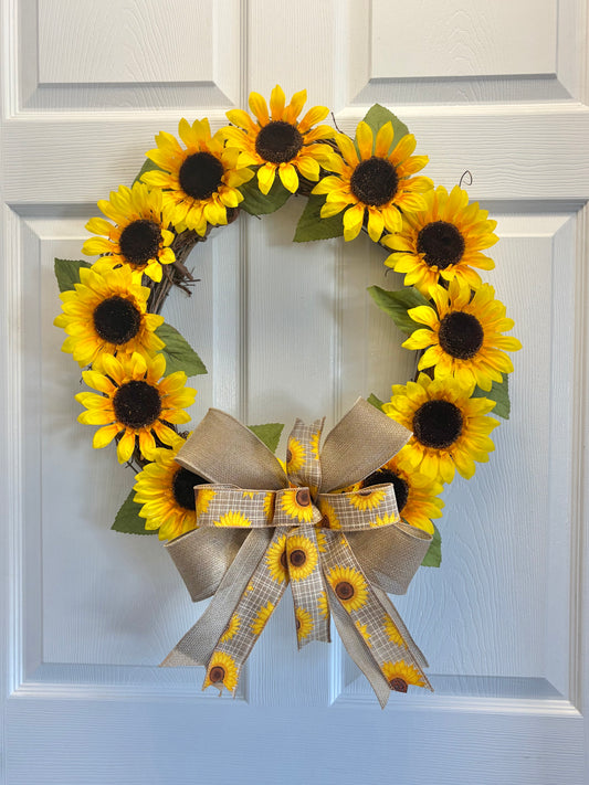 A sunflower wreath hanging on a white paneled door. The wreath features bright yellow artificial sunflowers with dark brown centers and green leaves, finished with a large tan ribbon bow and matching plaid ribbon accents.
