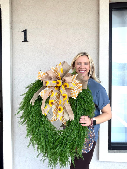 A smiling woman stands in front of a house door holding a large green wreath with a decorative bow. The bow is made from tan ribbon and yellow sunflower-patterned ribbon with green accents.