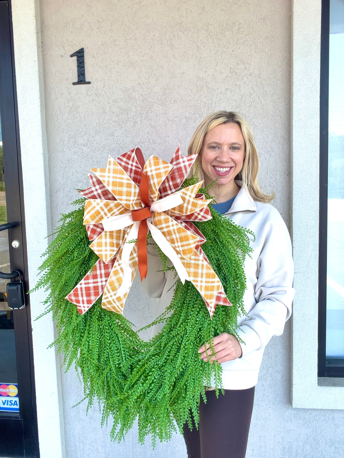A smiling woman stands in front of a beige house wall holding a green artificial bead grass wreath with a large plaid bow in orange, white, and rust tones.