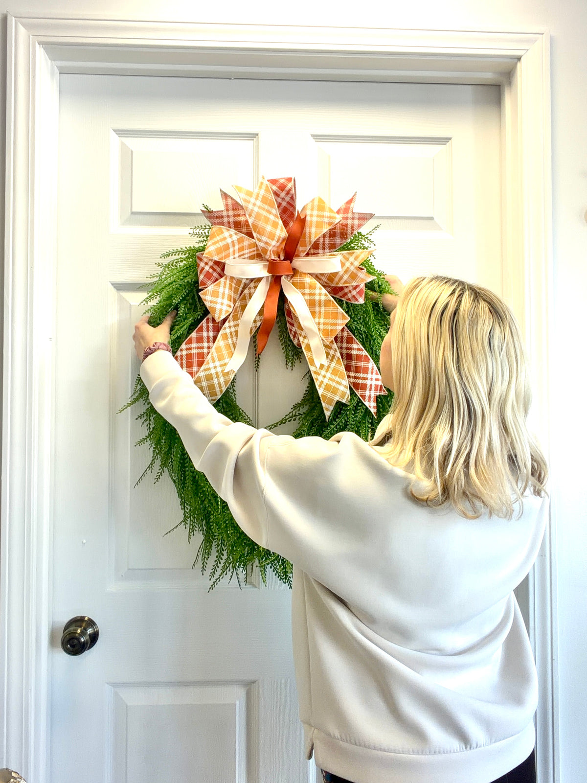 A woman with blonde hair, seen from behind, hangs a green bead grass wreath on a white paneled door. The wreath is decorated with a large orange and white plaid bow.