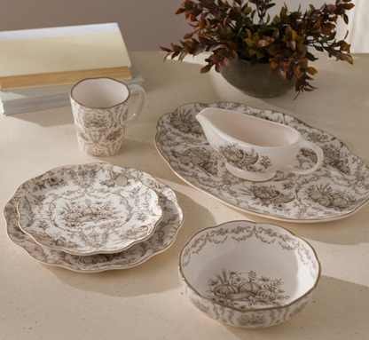 Elegant vintage-style dinnerware set with intricate botanical patterns in neutral tones, including plates, a gravy boat, and a mug, on a light-colored table with a plant in the background.