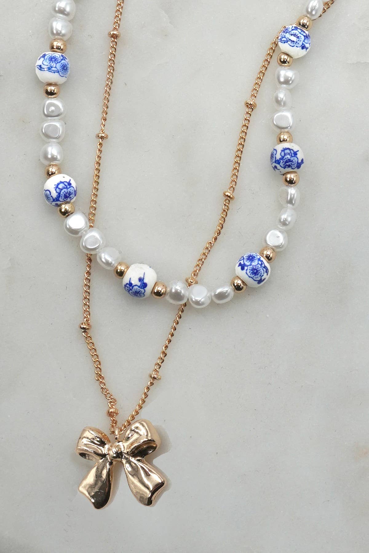 Layered necklace with gold chain with bow charm and white beads with blue toile pattern
