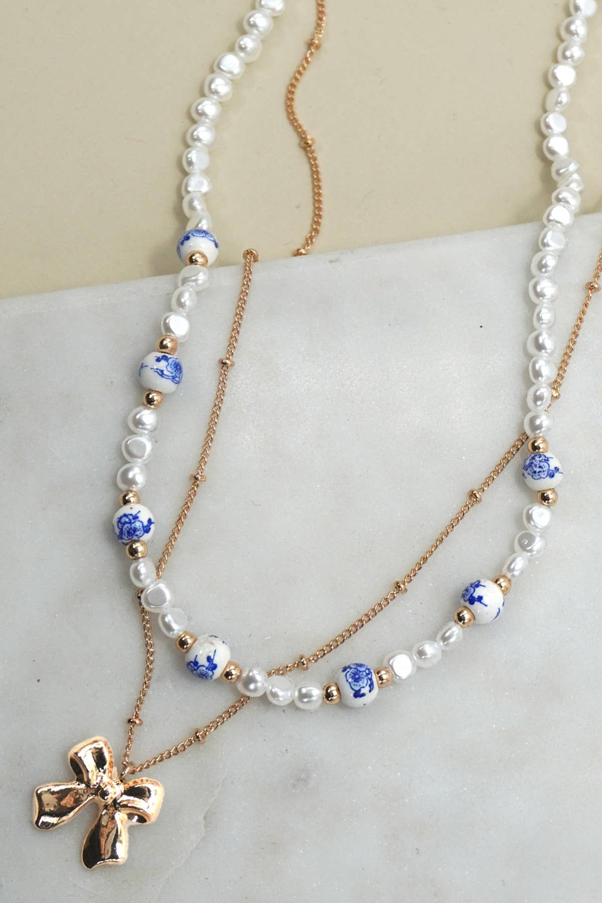 Layered necklace with gold chain with bow charm and white beads with blue toile pattern